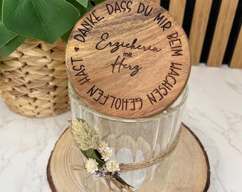 Könnte beinhalten: Ein durchsichtiger Glas-Kerzenhalter mit einem Holzdeckel. Der Deckel ist mit deutschem Text graviert. Der Kerzenhalter ist mit Bindfäden und Trockenblumen verziert und steht auf einer Holzscheibe.