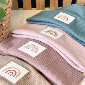 Könnte beinhalten: Vier gestrickte Beanies in verschiedenen Farben mit Regenbogen-Patches. Die Beanies sind übereinander gestapelt. Die Patches zeigen einen Regenbogen mit verschiedenen Farbstreifen und einer Vielzahl von Verzierungen.