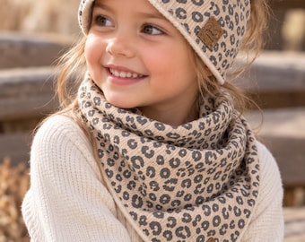 Op de afbeelding: Een beige en donkergrijze geribbelde hoofdband met luipaardprint, een gedraaide voorkant en een bijpassende driehoekige sjaal. Beide items zijn voorzien van een klein vierkant bruin lederachtig label met cursieve tekst.