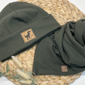 Może przedstawiać: Zestaw zielonego, żebrowanego beretu i pasującej szaliki z brązową skórzaną naszywką przedstawiającą grafikę dinozaura i napis "cool". Szalik ma brązową skórzaną naszywkę z napisem "Jovely Handmade".