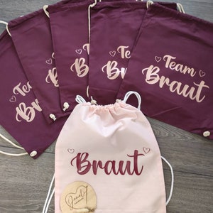 Może przedstawiać: Sześć burgundowych woreczków ze sznurkiem z napisem "Team Braut" i "Team Bride" złotymi literami. Jeden różowy woreczek ze sznurkiem z napisem "Braut" burgundowymi literami.