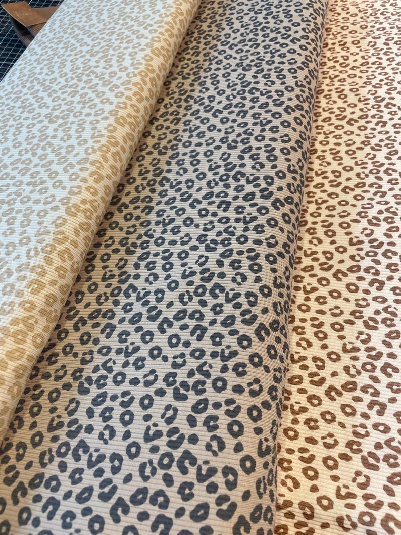 May include: Three rolls of fabric with a leopard print pattern. The fabric colors are beige, gray, and brown. The leopard spots are dark gray and brown. The fabric is ribbed.