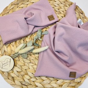 May include: A light purple ribbed knit headband and scarf set. The headband has a twisted design. Both pieces have a small, square, brown leather tag with the words "Lovely handmade" on them. The set is displayed on a woven, circular placemat.