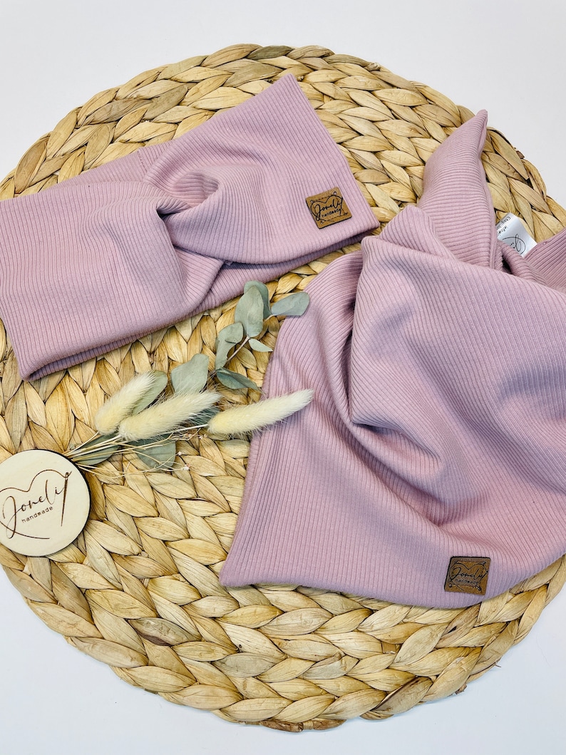 May include: A light purple ribbed knit headband and scarf set. The headband has a twisted design. Both pieces have a small, square, brown leather tag with the words "Lovely handmade" on them. The set is displayed on a woven, circular placemat.