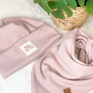 Può includere: Un berretto e una sciarpa abbinati di colore rosa a costine, con una grafica arcobaleno e il testo "Family First" su un'etichetta marrone.