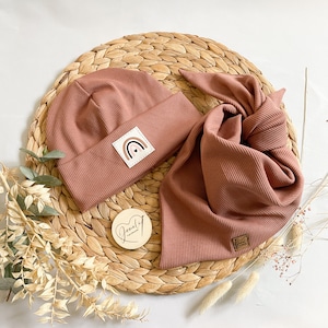 Peut inclure: Un ensemble de deux articles pour bébé de couleur rose poussiéreux. Un bonnet avec un patch arc-en-ciel et un bandana côtelé assorti. Les articles sont présentés sur un tapis de paille tressé.
