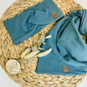 May include: Two pieces of teal-colored ribbed fabric, a headband and a scarf, are displayed on a woven wicker surface. Each piece has a small, square, brown leather tag. A small wooden disc with cursive text and dried flowers are also present.