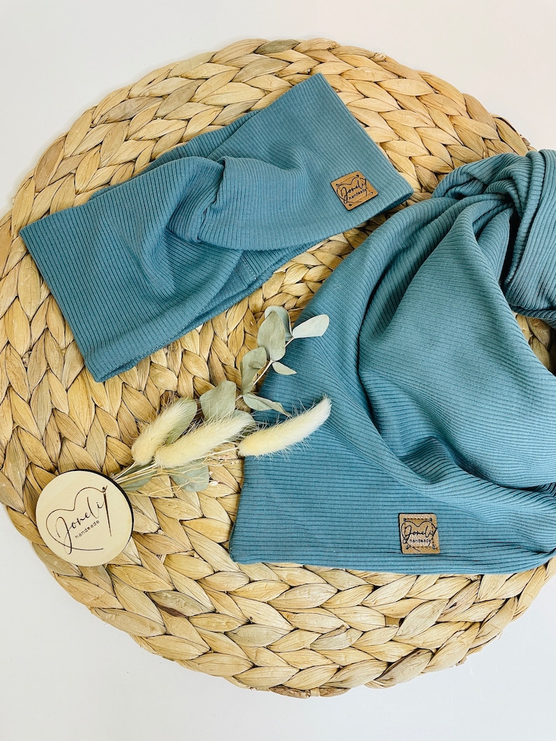 May include: Two pieces of teal-colored ribbed fabric, a headband and a scarf, are displayed on a woven wicker surface. Each piece has a small, square, brown leather tag. A small wooden disc with cursive text and dried flowers are also present.