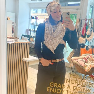 May include: A woman wearing a black long-sleeve shirt, dark jeans, and a brown belt. She is wearing a patterned scarf and headband. The mirror reflects the word "Hello" in gold script.