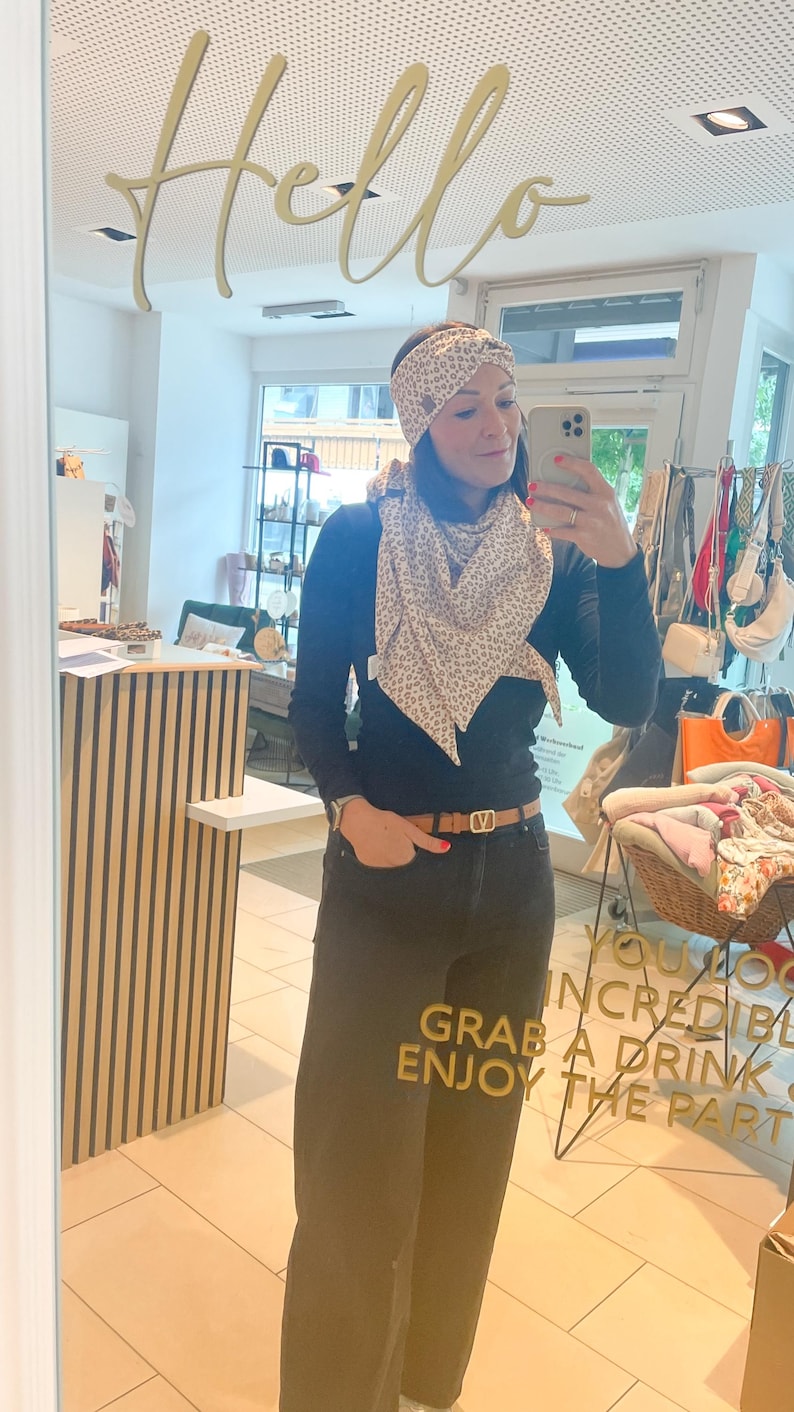 May include: A woman wearing a black long-sleeve shirt, dark jeans, and a brown belt. She is wearing a patterned scarf and headband. The mirror reflects the word "Hello" in gold script.