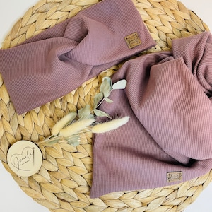 May include: A mauve-colored ribbed knit headband and scarf set. The headband features a twisted design, and both pieces have a small, square, brown label. The set is displayed on a woven, circular mat with dried floral accents and a wooden tag.