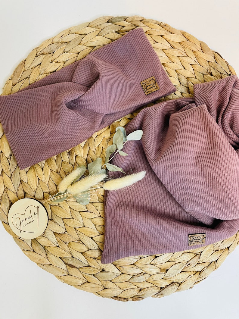May include: A mauve-colored ribbed knit headband and scarf set. The headband features a twisted design, and both pieces have a small, square, brown label. The set is displayed on a woven, circular mat with dried floral accents and a wooden tag.