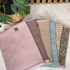 Pode incluir: Uma coleção de aquecedores de pescoço em várias cores, incluindo rosa, castanho, bege e azul, com uma opção de estampa de leopardo. Cada aquecedor de pescoço tem uma textura canelada e uma borda macia e contrastante. Um pequeno rótulo castanho é visível em alguns.