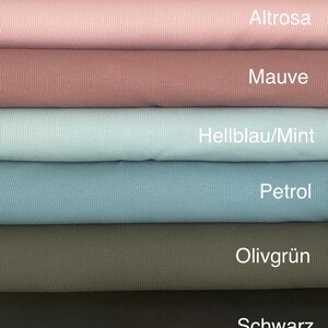 May include: A stack of various colored ribbed knit fabrics. Colors include beige, light pink, mauve, light blue, teal, olive green, and black. Each fabric is labeled with its color name. A small wooden tag with text and dried flowers are at the bottom.