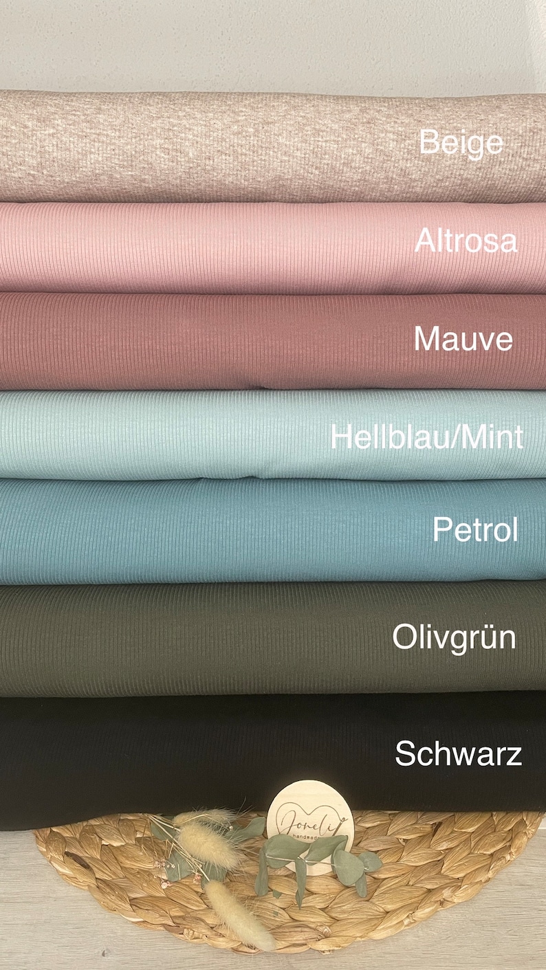 May include: A stack of various colored ribbed knit fabrics. Colors include beige, light pink, mauve, light blue, teal, olive green, and black. Each fabric is labeled with its color name. A small wooden tag with text and dried flowers are at the bottom.
