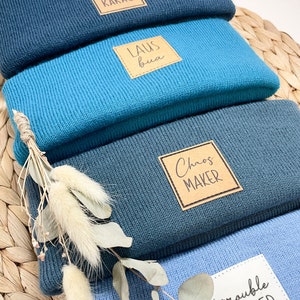 Könnte beinhalten: Ein Stapel von vier Strickmützen in Blautönen. Jede Mütze hat ein quadratisches Etikett mit den Texten: "ciao KAKAO", "LAUS bua", "Chaos MAKER" und "trouble MAKER". Getrocknete Blumen und Blätter sind daneben platziert.