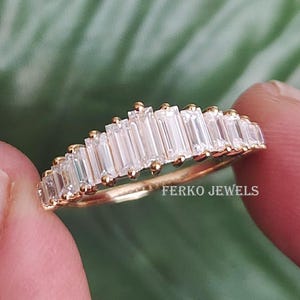 May include: A gold ring with a row of rectangular, clear gemstones. The ring is held between two fingers against a blurred green background. The text "FERKO JEWELS" is visible on the ring.