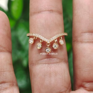 Op de afbeelding: Een goudkleurige ring met een V-vormig ontwerp, versierd met kleine, hangende ronde edelstenen. De ring heeft een rij kleine stenen langs de V-vorm en de merknaam "FERKO JEWELS". De ring wordt op een vinger getoond tegen een vage groene achtergrond.