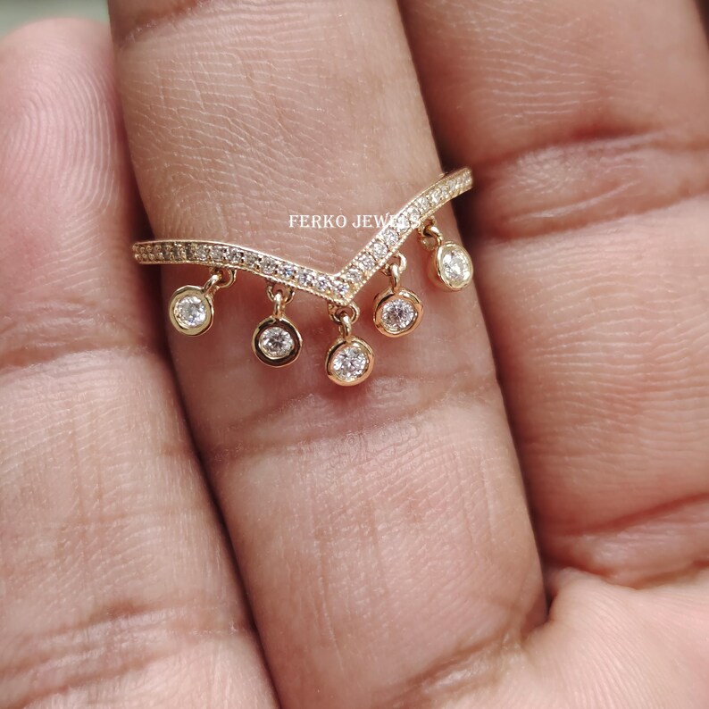 Op de afbeelding: Een goudkleurige ring met een V-vormig ontwerp. De ring is ingelegd met kleine, heldere stenen langs de bovenkant en heeft bungelende, ronde bedels met heldere stenen. De tekst "FERKO JEWEL" is zichtbaar.