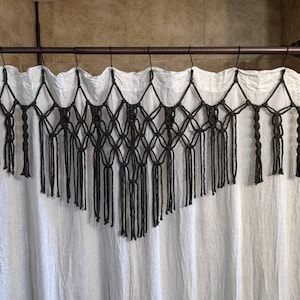 May include: A black macrame curtain valance with a diamond pattern and fringe. The valance is hanging on a white shower curtain.