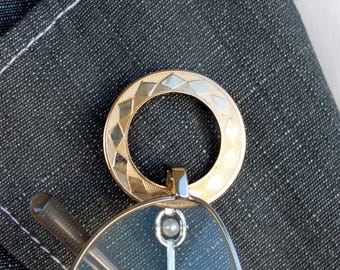 May include: A gold-toned circular brooch featuring a repeating diamond pattern along its outer edge. The brooch is attached to a pair of spectacles with a silver-coloured metal frame and clear lenses, resting against a dark grey textured fabric background.