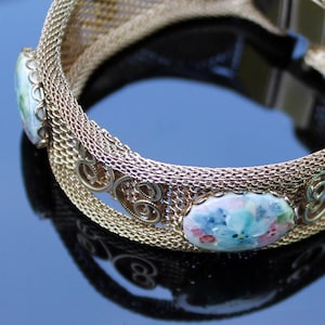 Könnte beinhalten: Ein goldenes Mesh-Armband mit zwei Porzellan-Blumenakzenten. Die Blumen sind in Blautönen, Rosa und Grün bemalt.