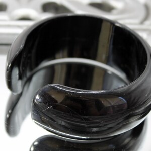 Könnte beinhalten: Ein breiter, polierter schwarzer Armreif mit einer glatten, abgerundeten Oberfläche und einem offenen C-förmigen Design. Das Material hat ein hochglänzendes Finish, das Licht reflektiert und helle Glanzlichter an den Kanten erzeugt.