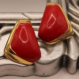 May include: A pair of red and gold clip-on earrings. The earrings are shaped like a triangle with rounded edges. The red part of the earrings is made of a smooth, shiny material. The gold part of the earrings is made of a shiny metal.