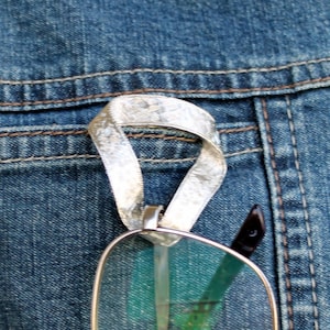 May include: A silver-colored metallic glasses holder clip featuring a looped, textured ribbon-like design. The clip is attached to a blue denim fabric surface, providing a secure way to hang eyewear when not in use by sliding the temple through the loop.