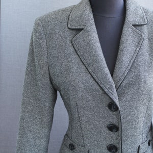 Puede incluir: Una chaqueta gris con ribetes y botones negros. La chaqueta tiene un cuello de muesca y un cierre de un solo botón.