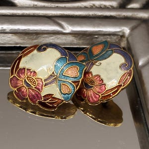 Può includere: Un paio di orecchini vintage con un design floreale e a farfalla. Gli orecchini presentano uno sfondo color crema con una farfalla blu e fiori rossi e viola. I bordi sono bordati in oro.