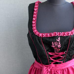 以下が含まれることがあります： ピンクのリボンで縁取られた黒いディルンドルで、ピンクの花の刺繍が施されています。ボディスはフロントにレースアップされており、スカートは無地のピンクです。