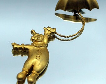 以下が含まれることがあります： 金色のピエロをテーマにしたブローチ。ブローチは、傘から吊り下げられたピエロの姿を特徴としています。ピエロは帽子とフリルカラーを着用しています。傘はピエロの上にあり、金色のチェーンでつながっています。
