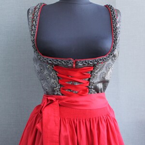 May include: A traditional dirndl dress, featuring a gray bodice with floral patterns and red trim, a red ribbon corset, and a matching red skirt. The dress is displayed on a mannequin against a gray backdrop.