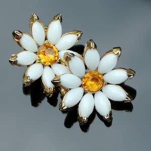 Könnte beinhalten: Ein Paar goldfarbene Ohrringe in Form von Gänseblümchen mit weißen Emailleblüten und orangefarbenen Strasssteinen in der Mitte.