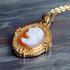 Op de afbeelding: Een goudkleurige camee hanger ketting. De ovale hanger heeft een wit profiel tegen een oranje achtergrond, omlijst door een getextureerde gouden rand en kleine kristallen. De ketting is ook goudkleurig.