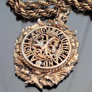 Puede incluir: Un collar con un colgante de tono dorado con un gran colgante adornado con un escudo con las palabras "ARGYLL AND SUTHERLAND" alrededor del borde. El colgante está decorado con motivos de cardo y una corona estilizada.