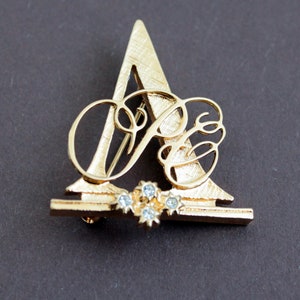 Puede incluir: Broche dorada con una letra 'A' estilizada con una 'B' más pequeña en su interior, acentuada con diamantes de imitación transparentes.