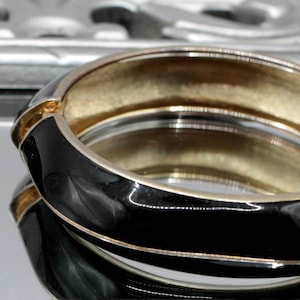 May include: A black enamel bangle bracelet with a gold-toned metal band. The bracelet is open and has a simple design.