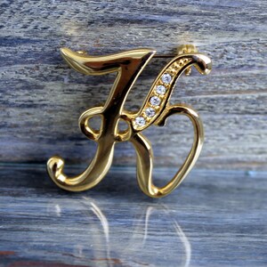 May include: A gold-tone letter K brooch with clear rhinestones. The letter is in a cursive font and the rhinestones are set in a row along the top of the letter.