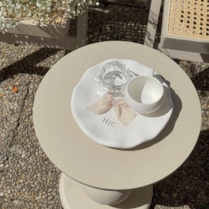 May include: A small, round, beige side table with a white plate, a clear glass of water, and a white cup. The plate has a bow and the initials "H|C". The table is outdoors, with a stone surface.