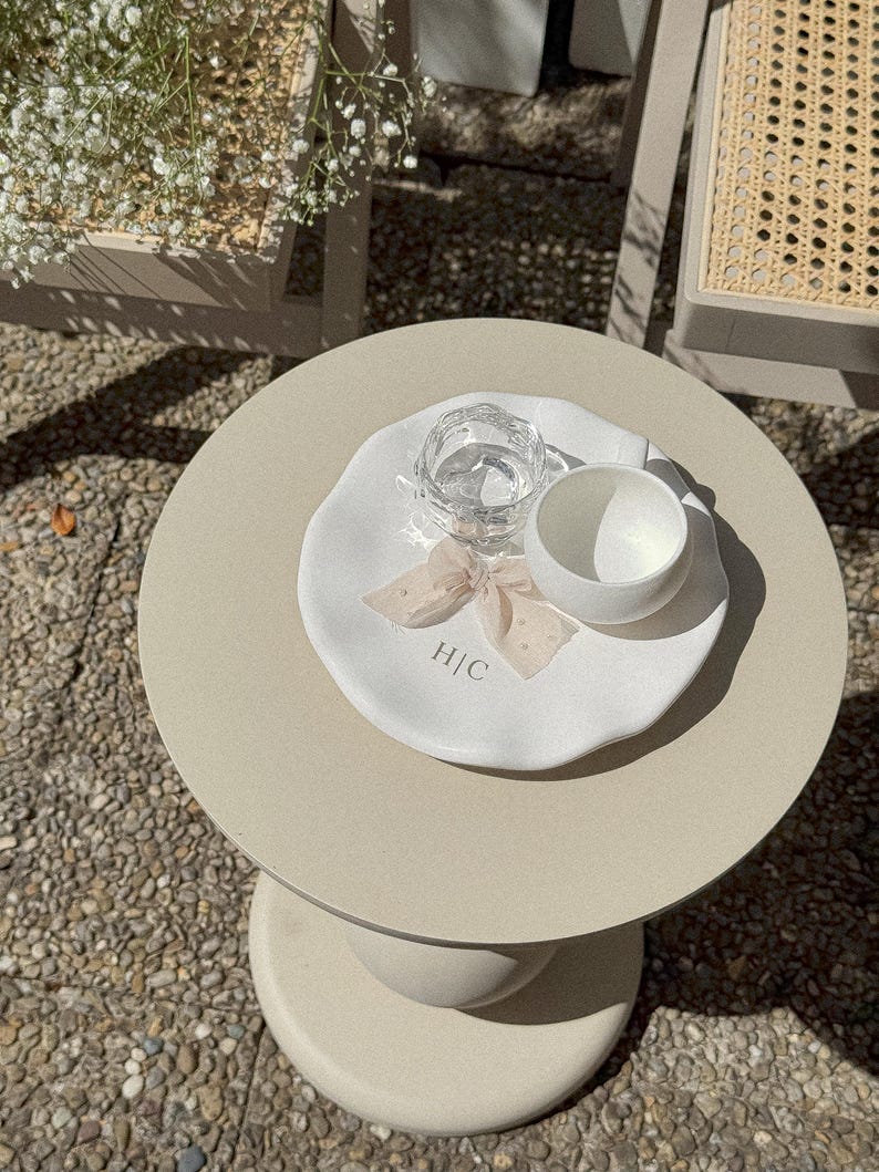 May include: A small, round, beige side table with a white plate, a clear glass of water, and a white cup. The plate has a bow and the initials "H|C". The table is outdoors, with a stone surface.