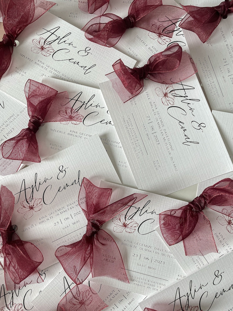 May include: Burgundy red satin ribbon bows tied around white wedding invitations with the text "Aylin & Cemal" and the date "23|06|2023".