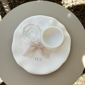 May include: A white ceramic tray with a clear glass dish, a white teacup, and a beige bow with pearl accents. The tray has the initials "H|C" printed on it. The items are arranged on a round, light gray table.