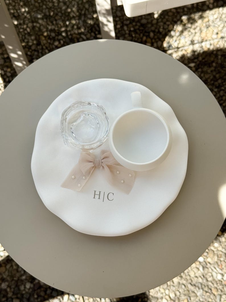 May include: A white ceramic tray with a clear glass dish, a white teacup, and a beige bow with pearl accents. The tray has the initials "H|C" printed on it. The items are arranged on a round, light gray table.