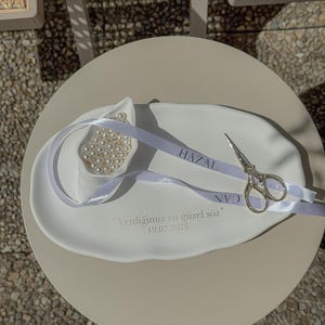 May include: A white ceramic dish with a small bowl filled with pearls, a silver ribbon with text, and small silver scissors. The dish has text and the date 19.07.2025. The items are on a round, light-colored table.