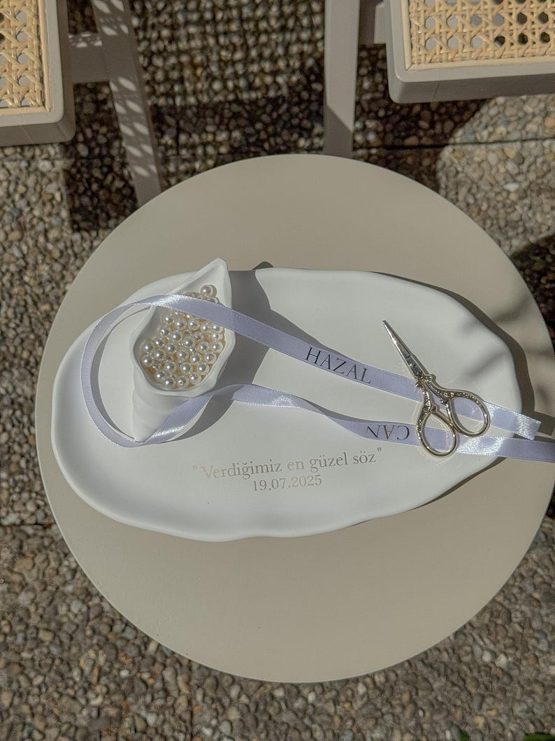 May include: A white ceramic dish with a small bowl filled with pearls, a silver ribbon with text, and small silver scissors. The dish has text and the date 19.07.2025. The items are on a round, light-colored table.