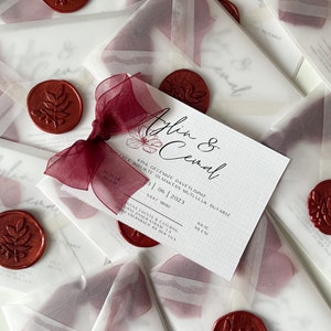 May include: A white invitation with a red wax seal and a burgundy ribbon. The invitation reads "Aplin & Cemal" and includes the date "23|06|2023" and the time "1500".