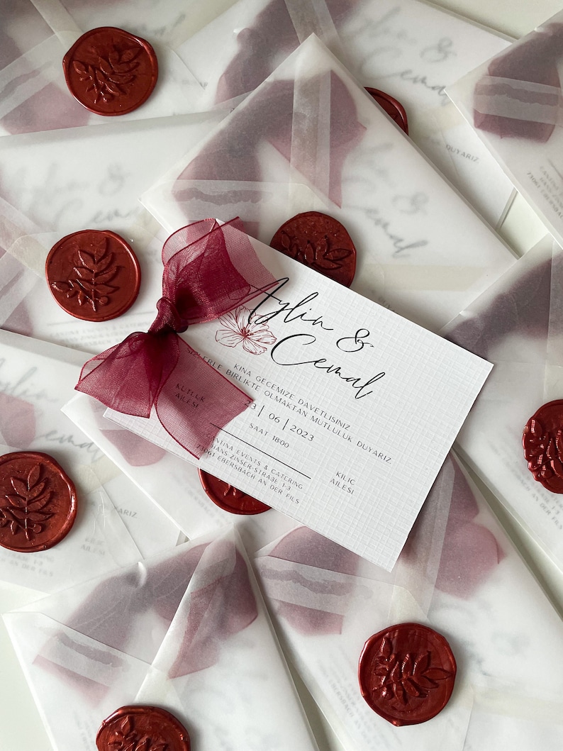 May include: A white invitation with a red wax seal and a burgundy ribbon. The invitation reads "Aplin & Cemal" and includes the date "23|06|2023" and the time "1500".