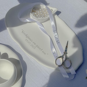 May include: A white ceramic dish with a small shell-shaped compartment filled with pearls. A white ribbon with the name "HAZAL" and "CAN" is draped across the dish, along with small silver scissors. The dish has text that reads "Verdiğimiz en g&uuml;zel s&ouml;z" and the date "19.07.2023".